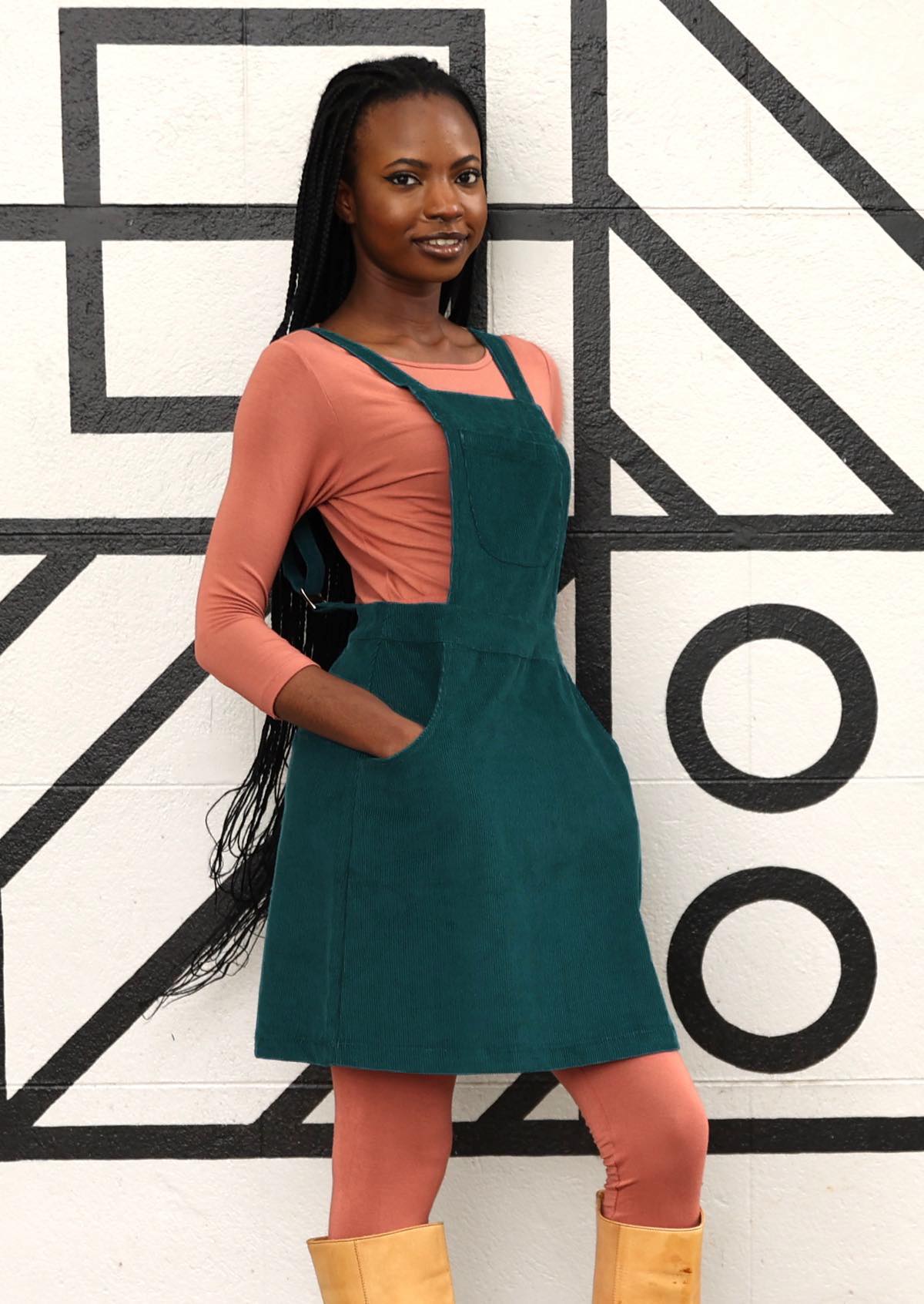 woman wearing teal corduroy pinafore over pink top, pink leggings and tan boots