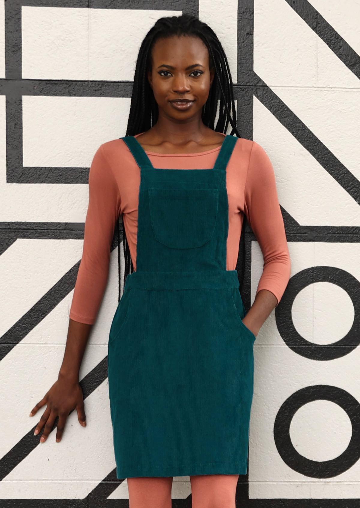 woman wearing teal corduroy pinafore over pink top, pink leggings and tan boots with hand in pocket