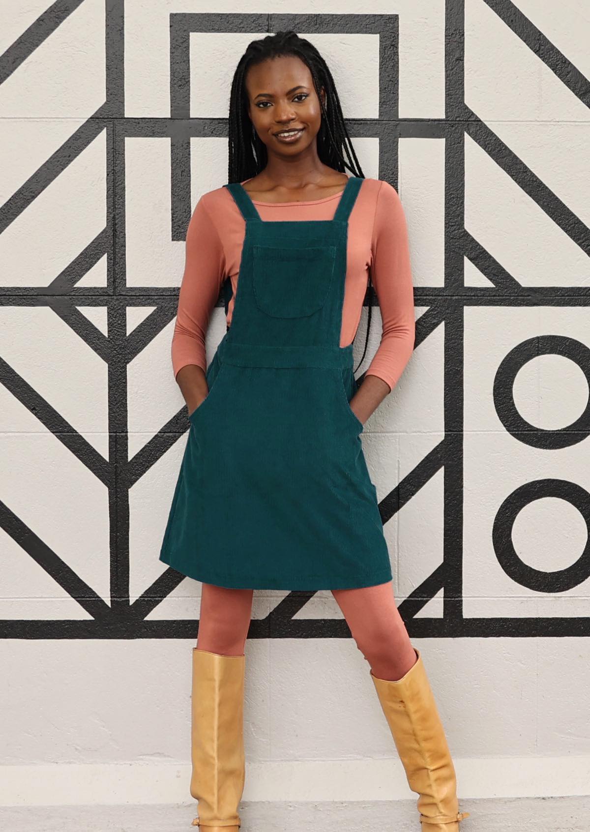 woman wearing teal corduroy pinafore over pink top, pink leggings and tan boots in front of black and white mural