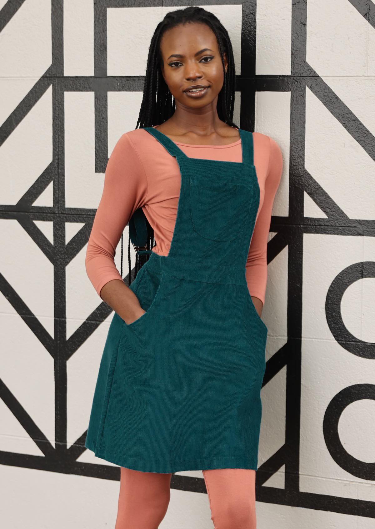 woman wearing teal corduroy pinafore over pink top, pink leggings leaning against wall