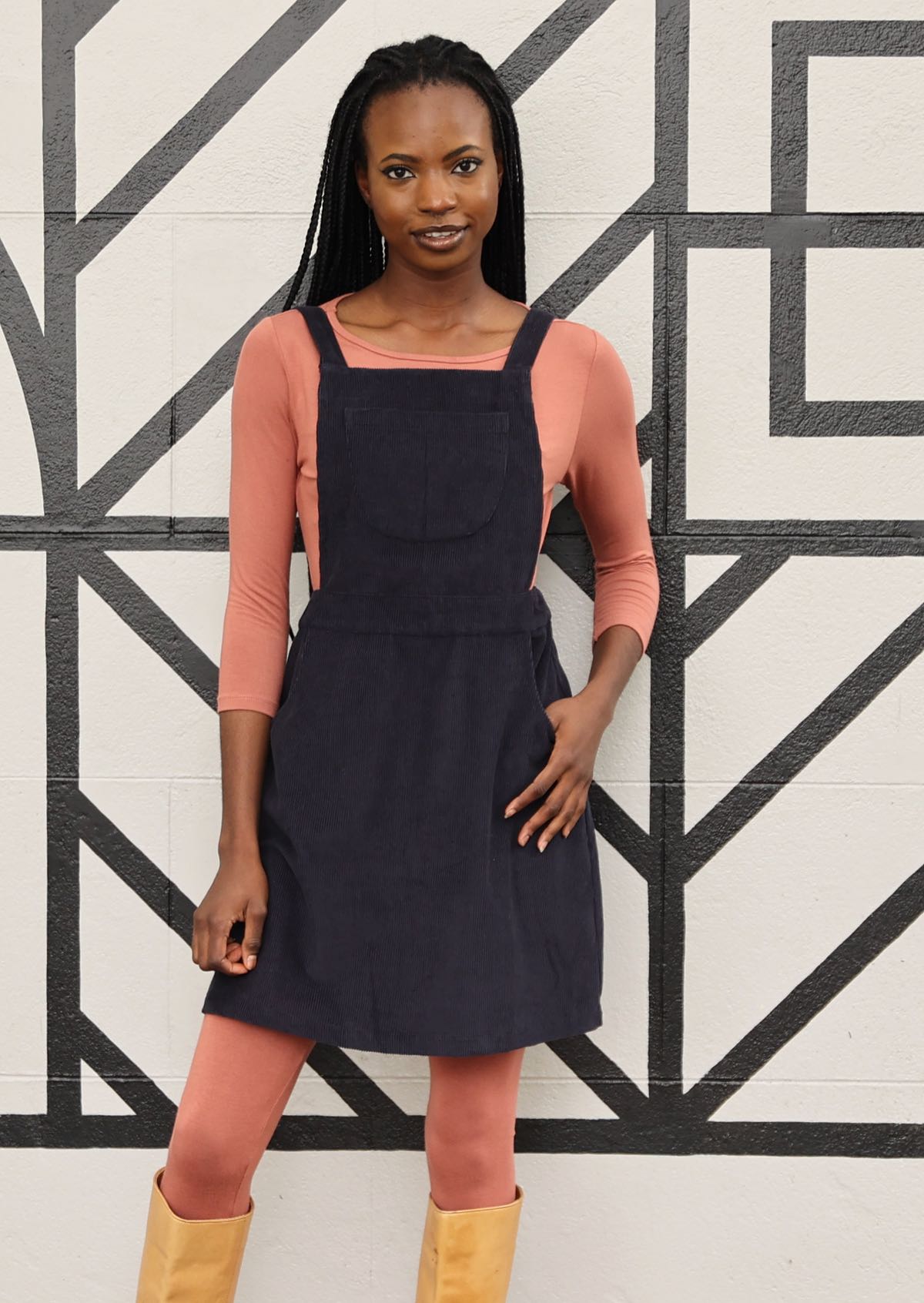 woman wearing dark grey cotton corduroy pinafore over pink top and pink leggings in front of black and white mural