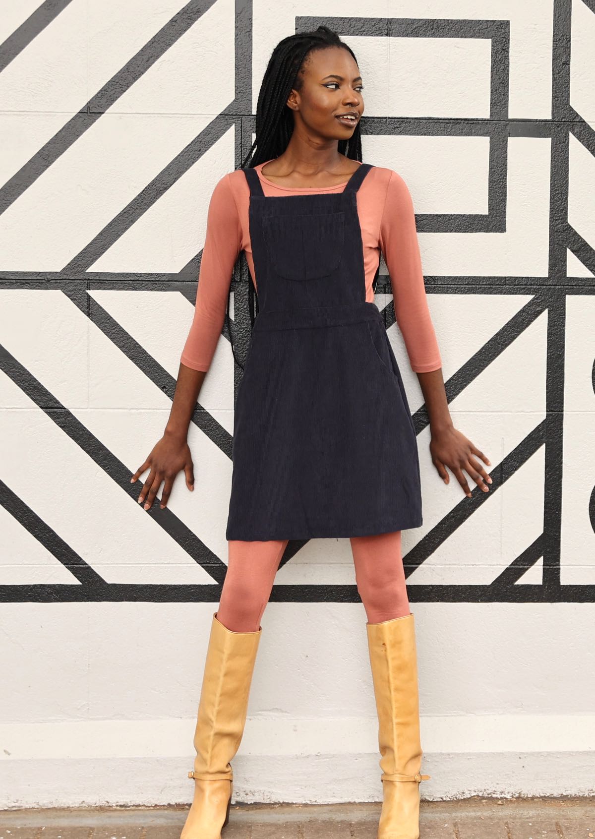 woman wearing dark grey cotton corduroy pinafore over pink top and pink leggings with tan boots