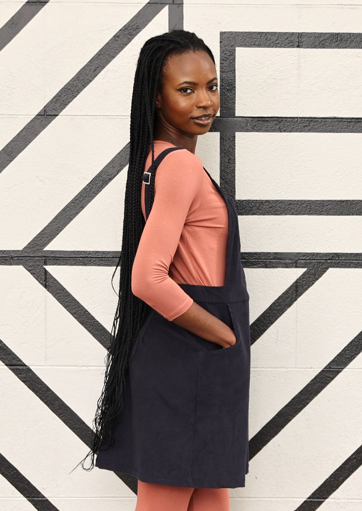 woman wearing dark grey cotton corduroy pinafore over pink top and pink leggings with hands in pockets
