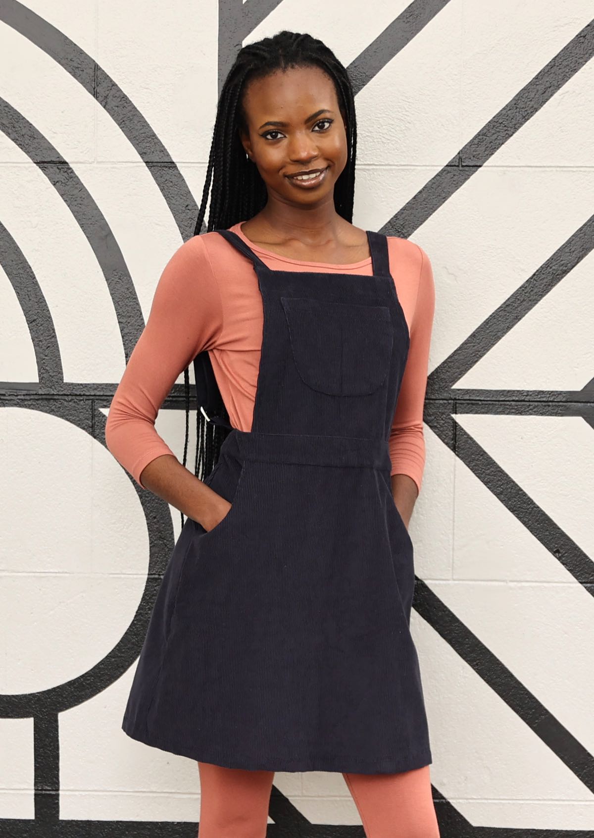woman wearing dark grey cotton corduroy pinafore over pink top and pink leggings