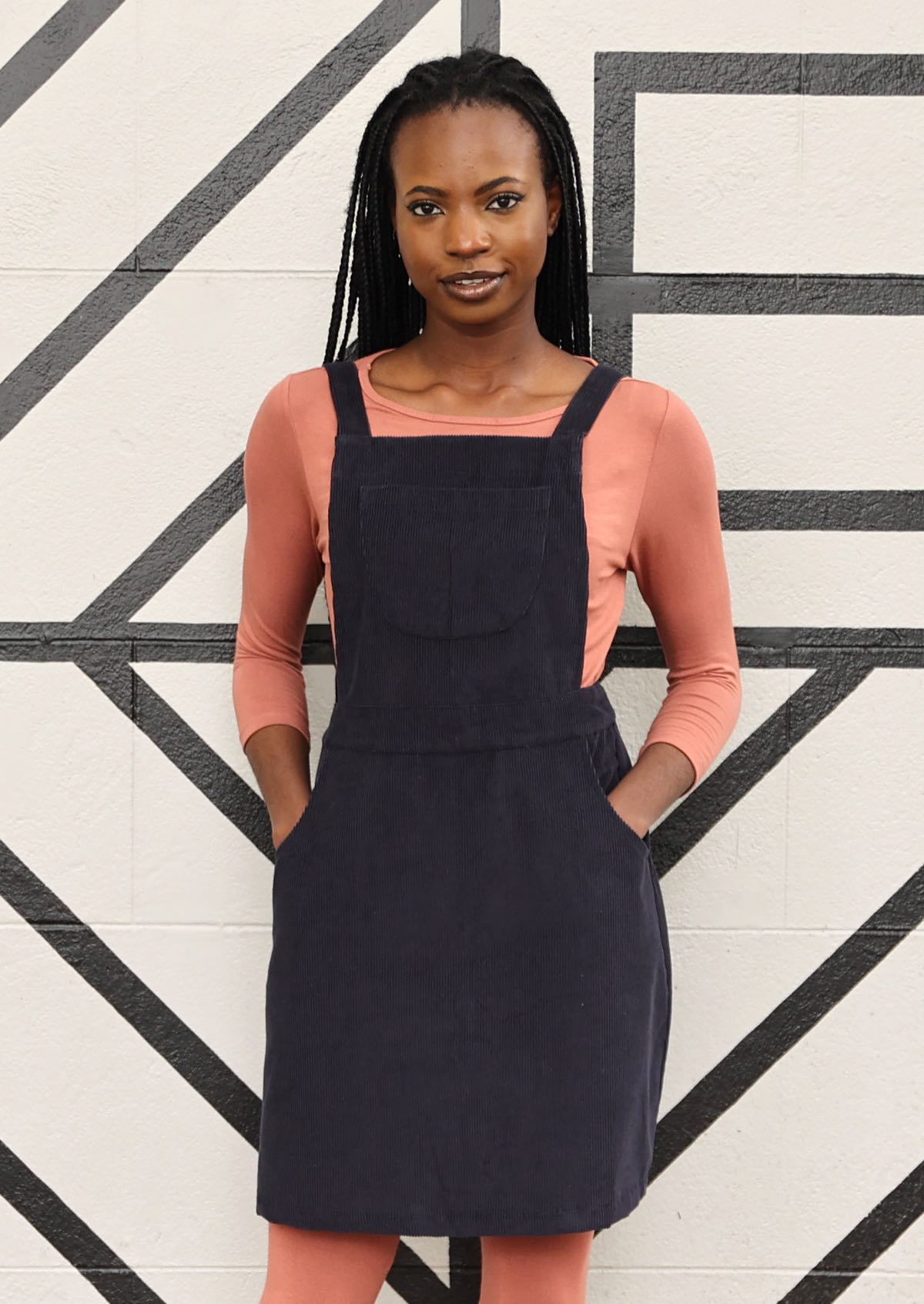 woman wearing dark grey cotton corduroy pinafore over pink top and pink leggings with hands in pockets
