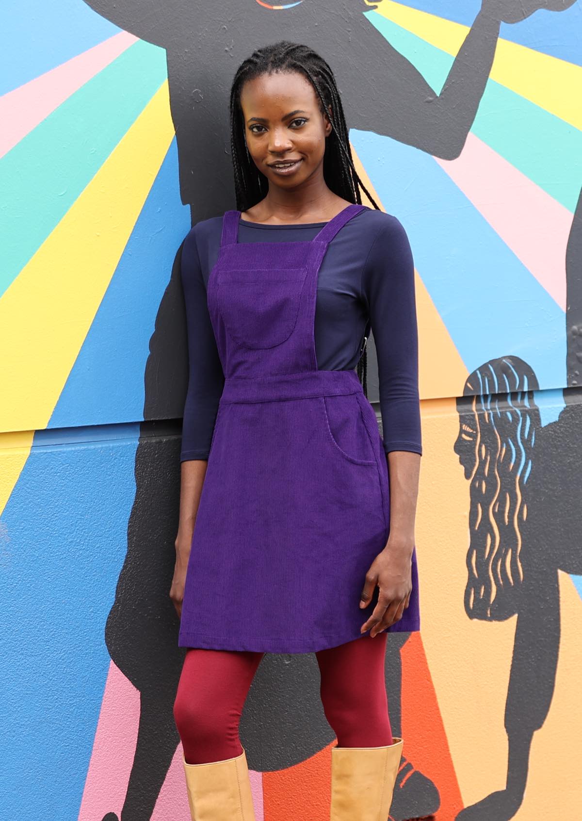woman wearing purple cotton corduroy pinafore over navy blue top and maroon leggings leaning against bright painted wall
