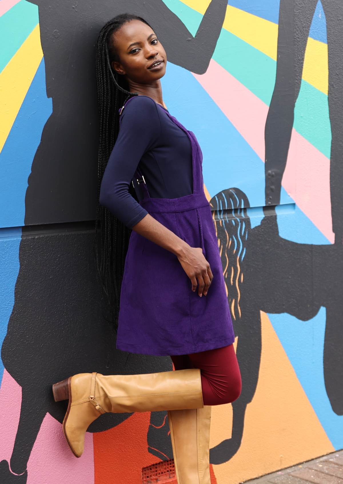 woman wearing purple cotton corduroy pinafore over navy blue top and maroon leggings and long tan boots