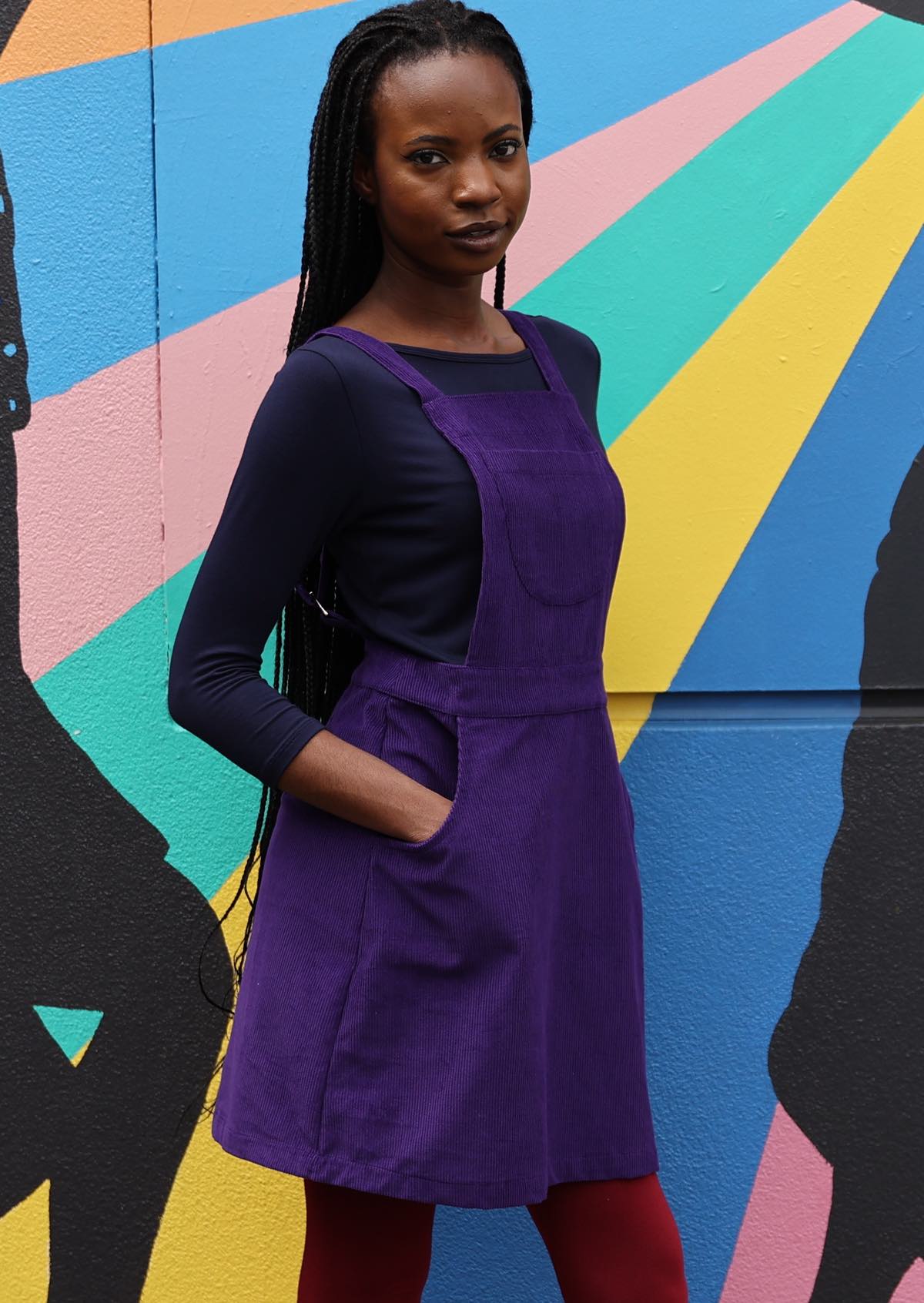 woman wearing purple cotton corduroy pinafore over navy blue top and maroon leggings