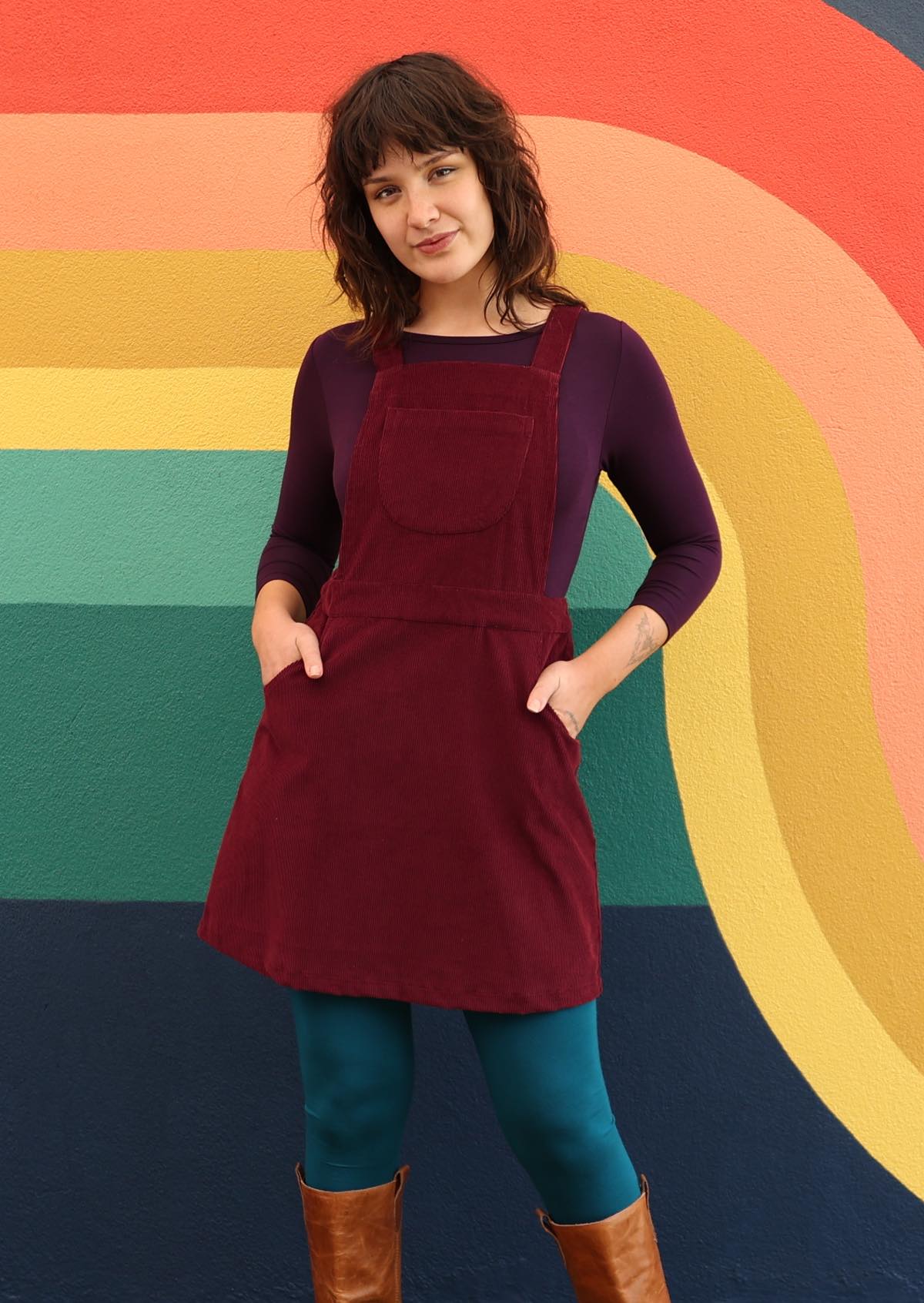 woman wearing maroon cotton corduroy pinafore over purple top and teal leggings with hands in pockets