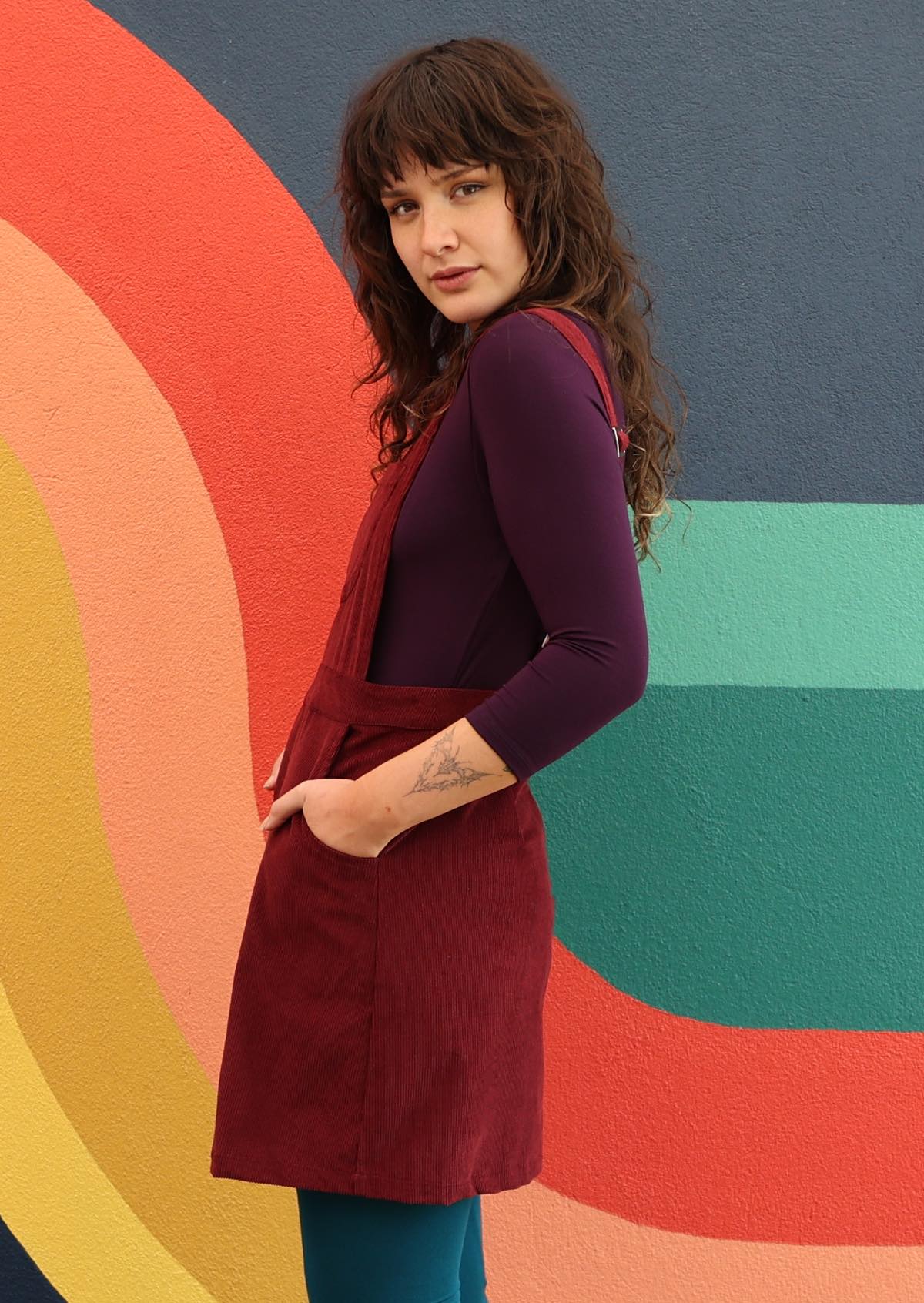 woman wearing maroon cotton corduroy pinafore over purple top and teal leggings side view