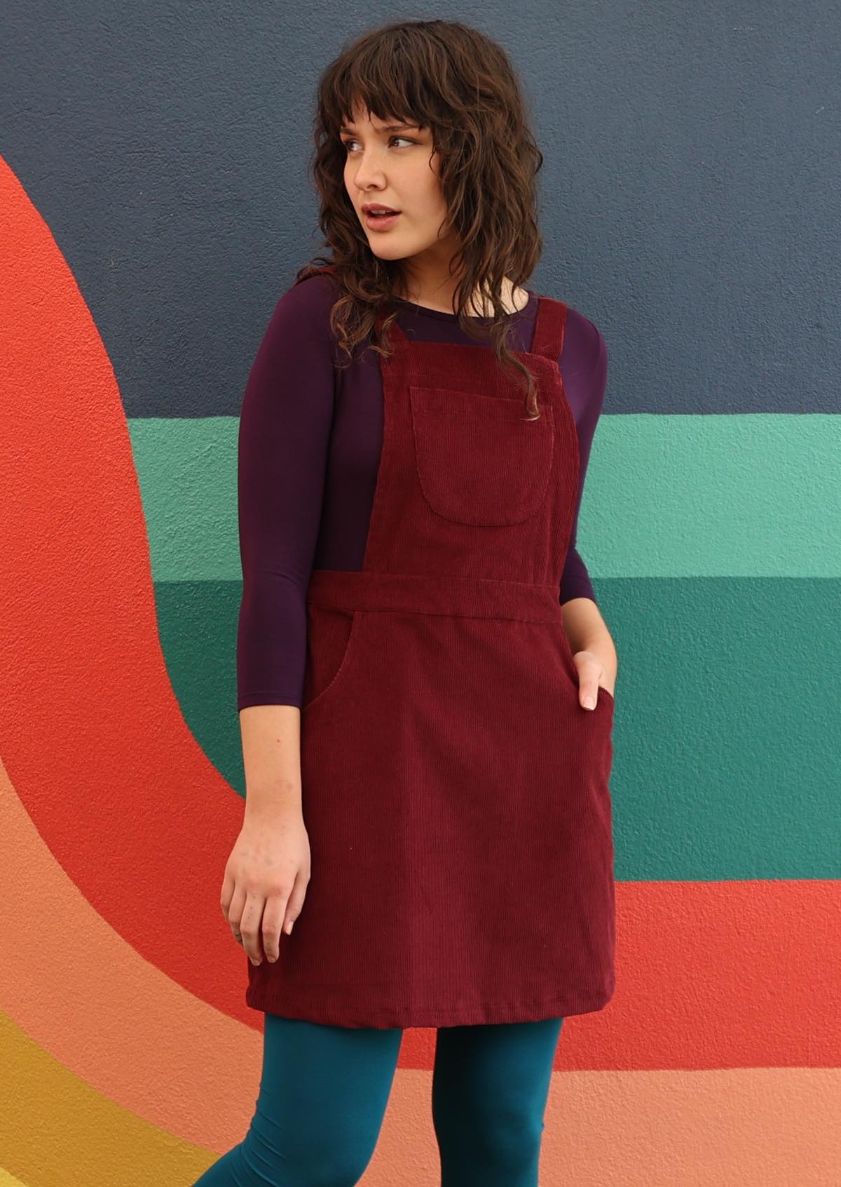woman wearing maroon cotton corduroy pinafore over purple top and teal leggings