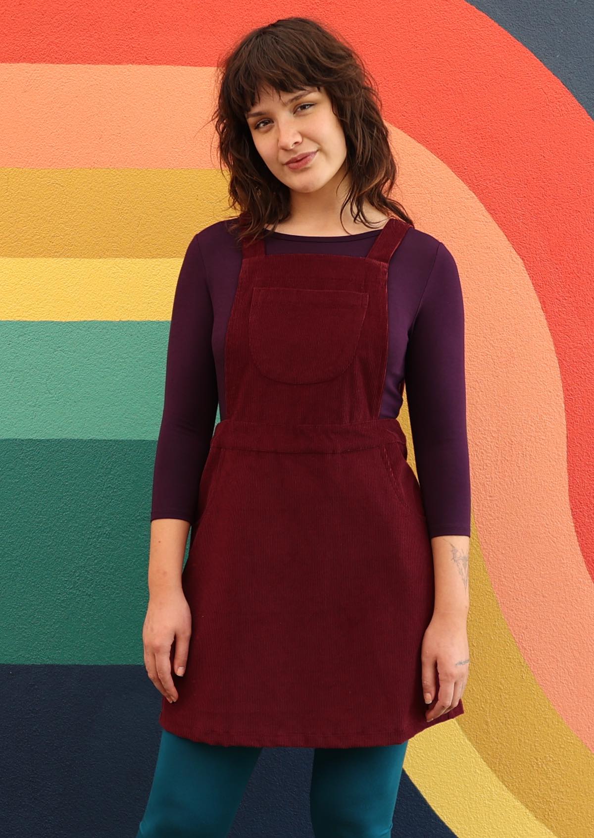 woman wearing maroon cotton corduroy pinafore over purple top and teal leggings in front of bright mural