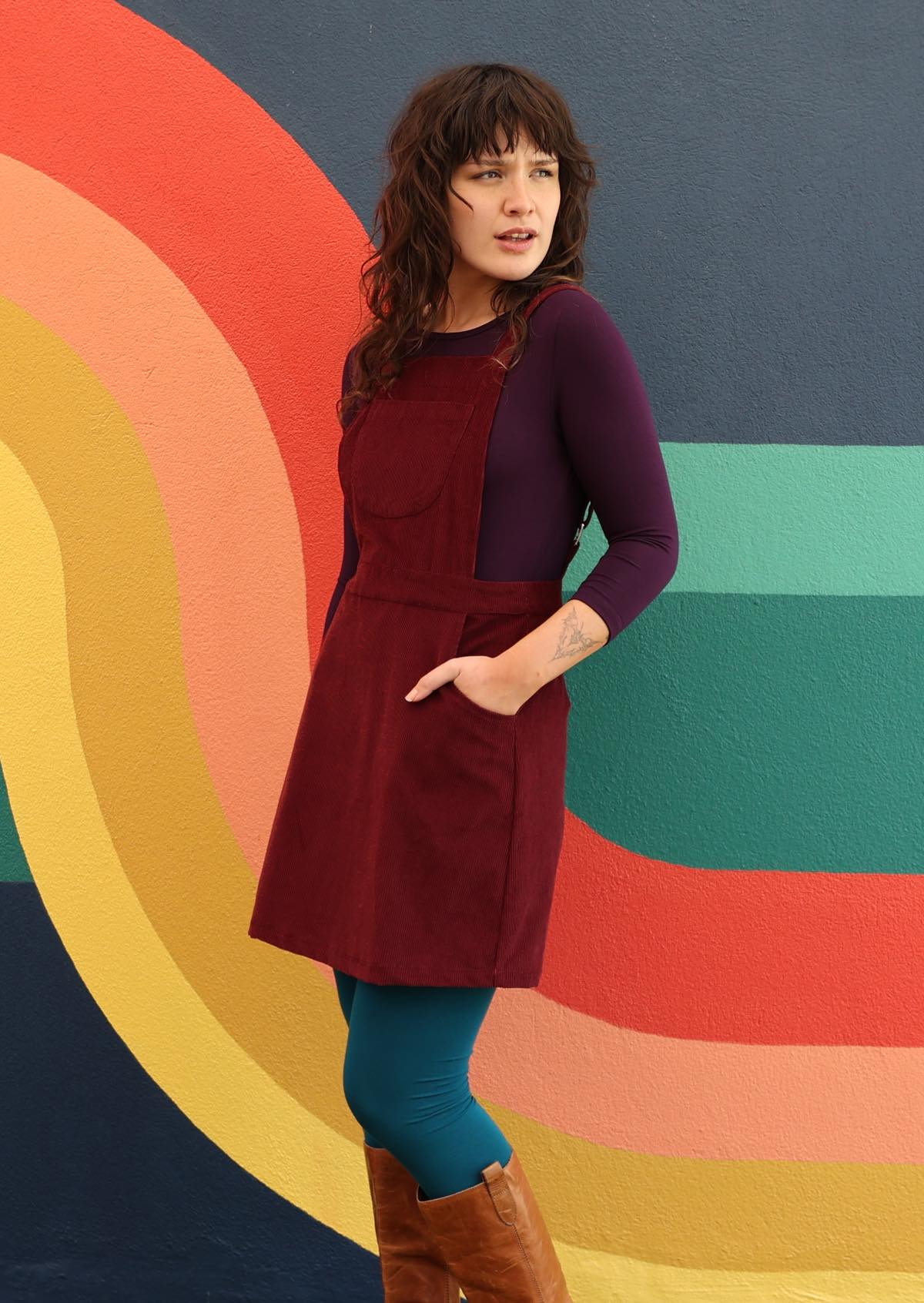 woman wearing maroon cotton corduroy pinafore over purple top and teal leggings in front of 70's style mural