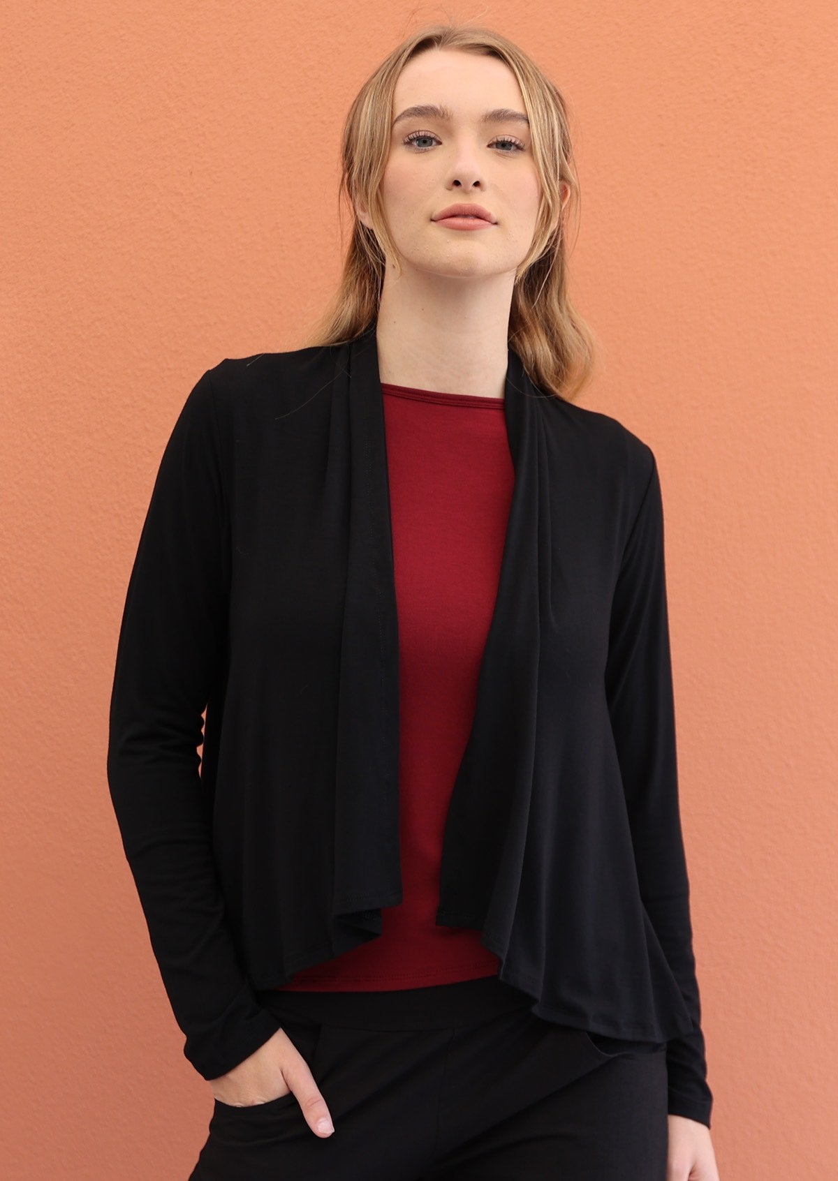 Model wears black long sleeve stretch rayon cardi with a waterfall front hem that is cropped at the front, exposing maroon top underneath