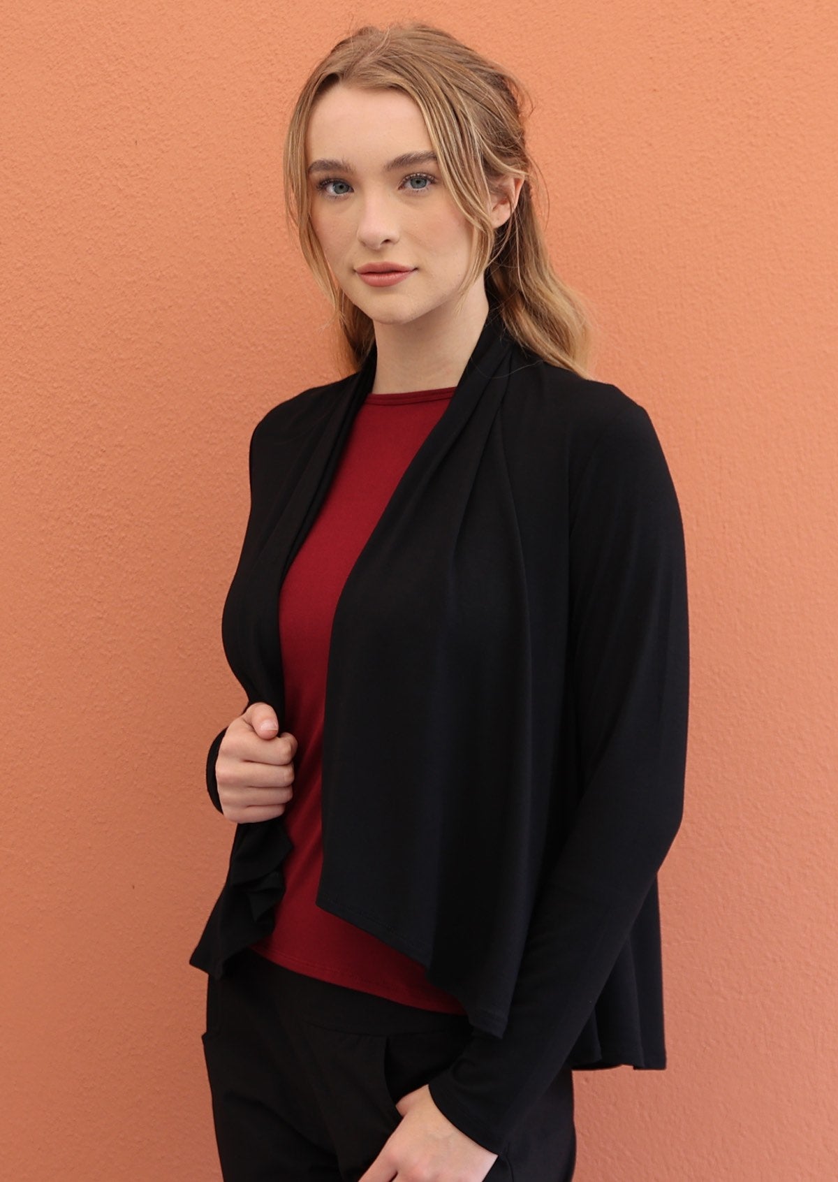 Woman wears black long sleeve stretch rayon cardi with a waterfall front hem, that reveals maroon top underneath
