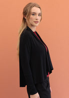 Woman stands side on in black long sleeve stretch rayon cardi with a waterfall front hem that is cropped at the front whilst back is longer