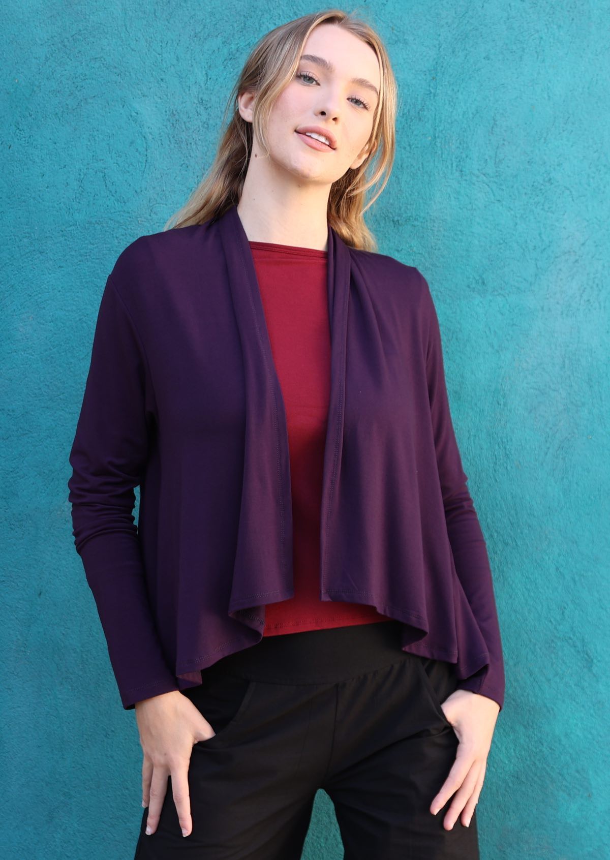 Woman wears long sleeve stretch rayon cardi in dark purple, with waterfall front hem that exposes maroon top underneath