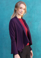 Woman stands side on in long sleeve stretch rayon cardi in dark purple, with waterfall front hem that exposes maroon top underneath