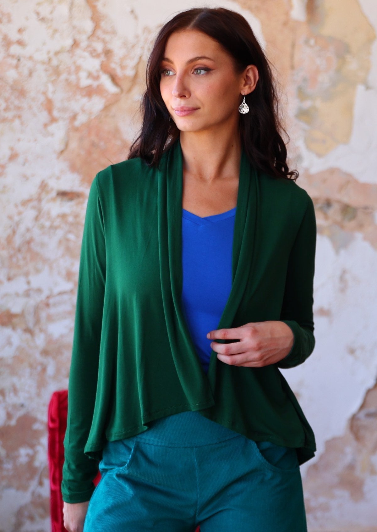Woman wears forest green long sleeve stretch rayon cardi with a waterfall front hem, that reveals bright blue top underneath