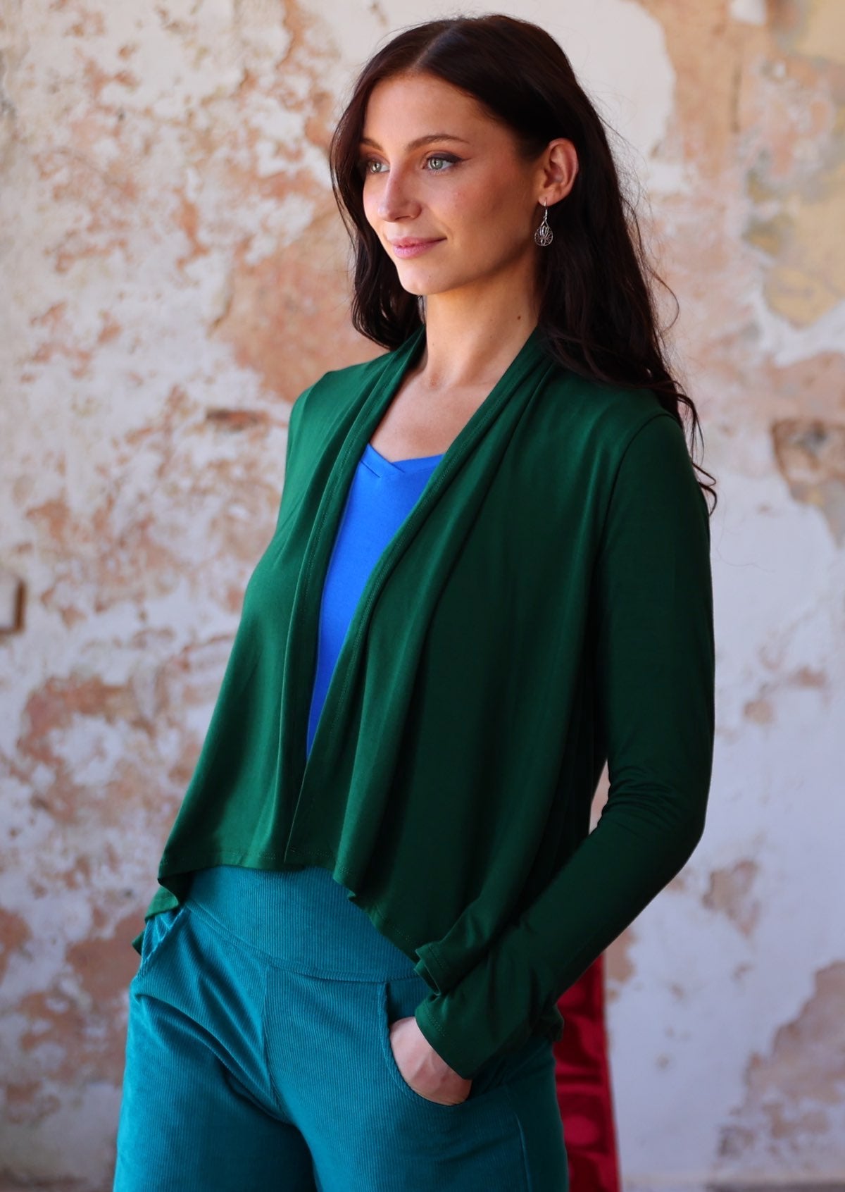 Woman stands side on in forest green long sleeve stretch rayon cardi with a waterfall front hem that is cropped at the front whilst back is longer