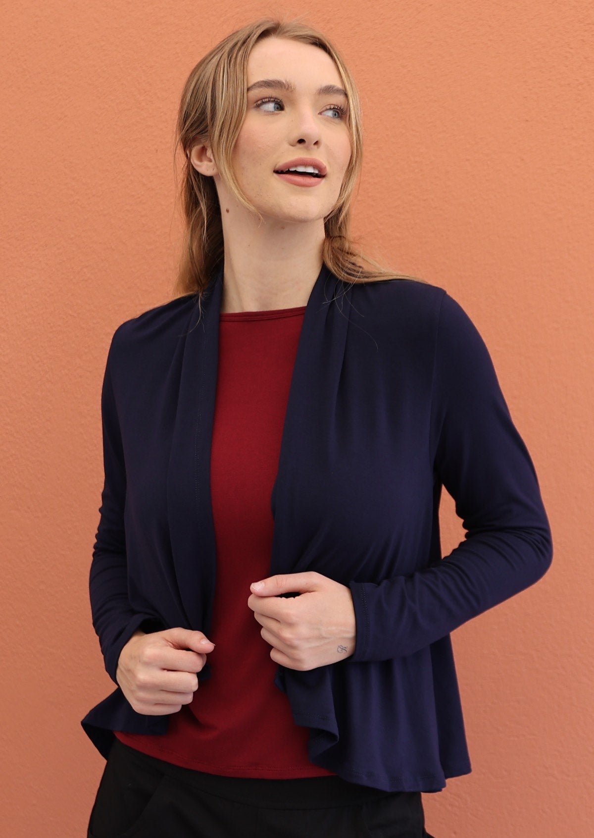 Woman wears navy blue long sleeve stretch rayon cardi with waterfall front hem that exposes maroon top and black pants underneath