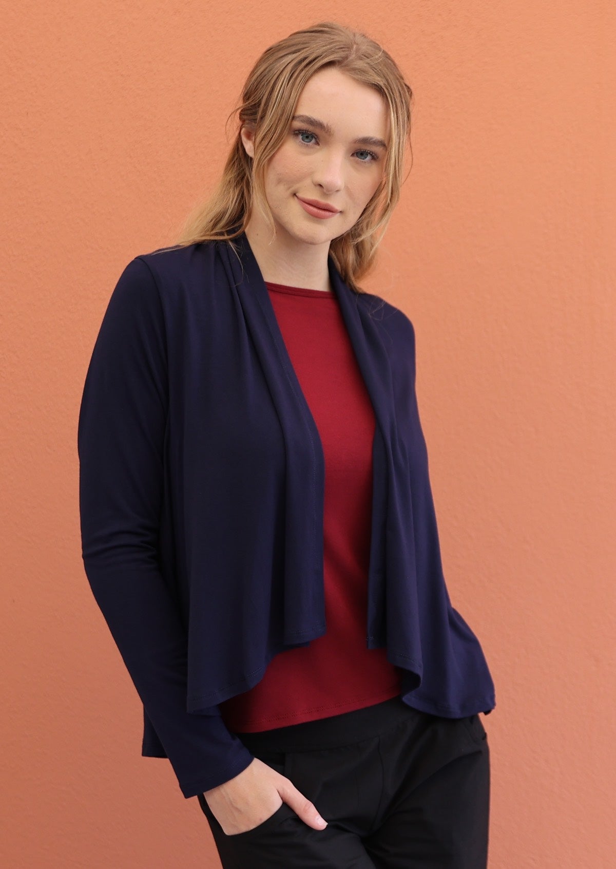 Woman poses in navy blue long sleeve stretch rayon cardi with waterfall front hem, with hands in pockets of black pants
