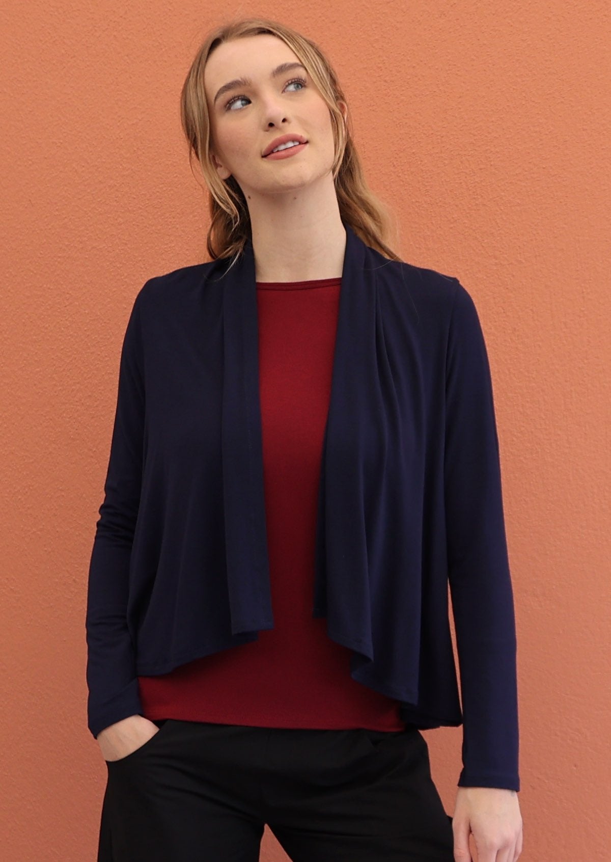 Model wears navy blue long sleeve stretch rayon cardi with waterfall front hem that exposes maroon top and black pants underneath