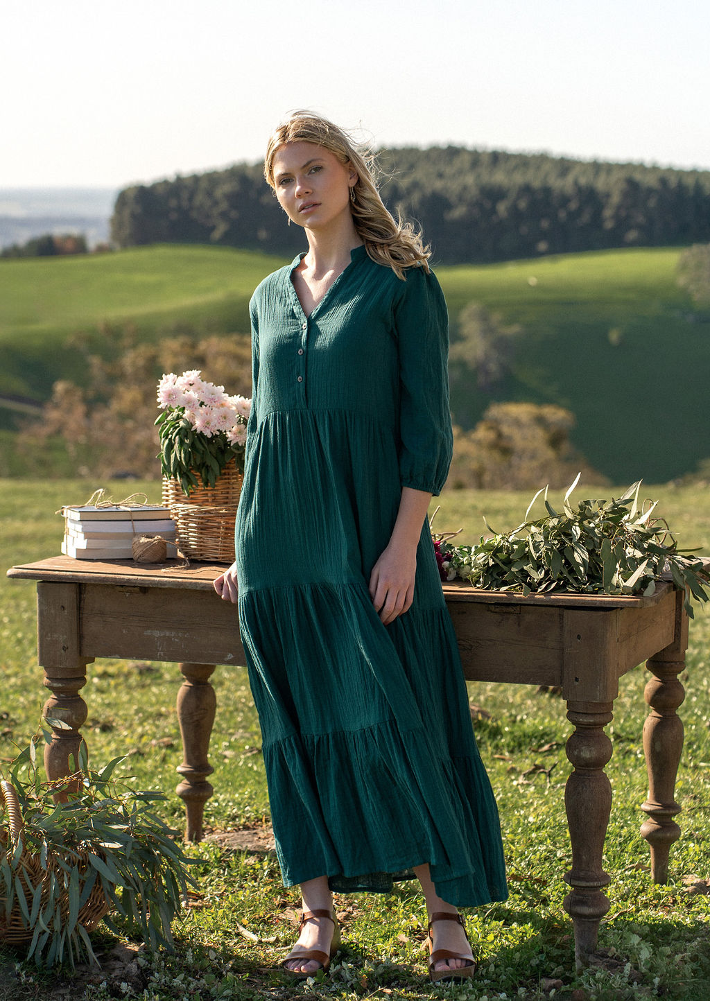 Women in deep teal cotton maxi dress in the country in front of old rustic table covered in flowers and gum leaves