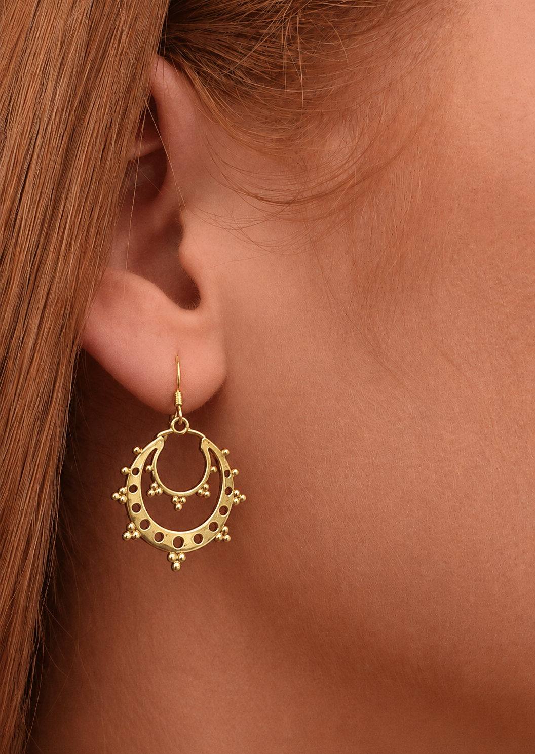 Close-up of a woman wearing delicate gold coloured crescent hoop earrings that have open cut-out detailing and mini bead accents.