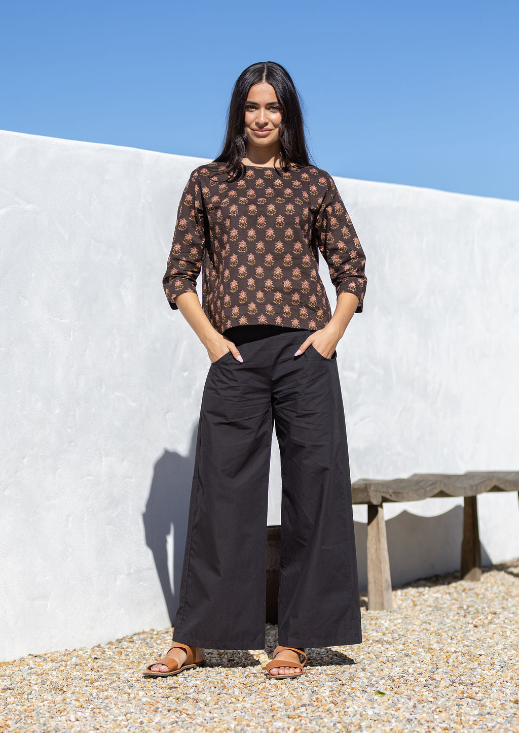 Woman wearing a brown cotton patterned top and black pants standing against a white wall with blue sky.