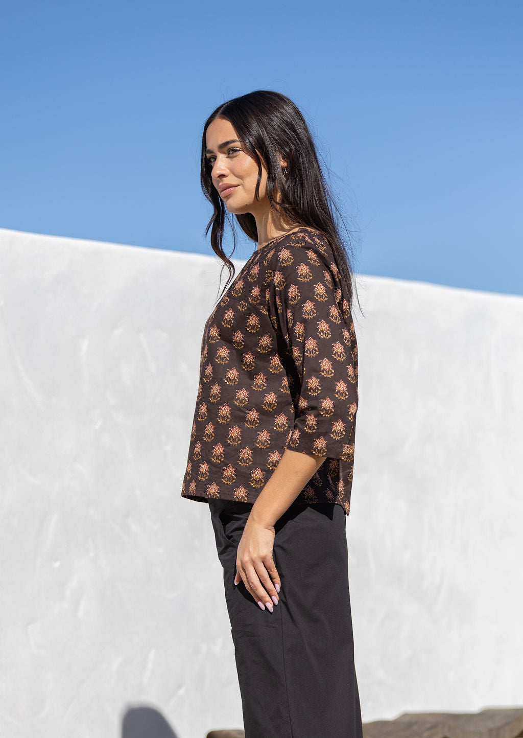 Woman wearing a brown Indian cotton patterned top against a white wall and blue sky