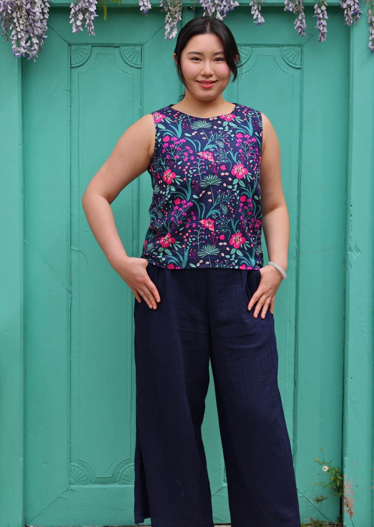 Model standing in front of aqua door with thumbs in pockets 100 percent cotton pink and purple florals on a dark blue base sleeveless top