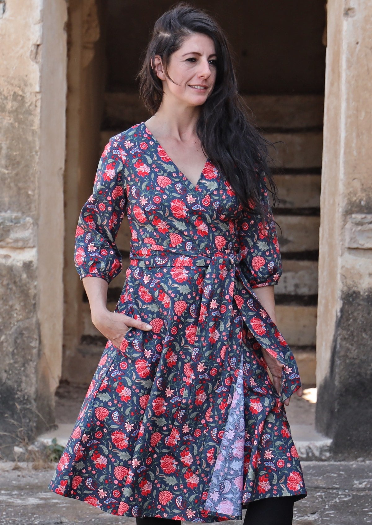 Woman in black hair, wearing over the knee length wrap dress in navy blue based cotton print and a hand in pocket.