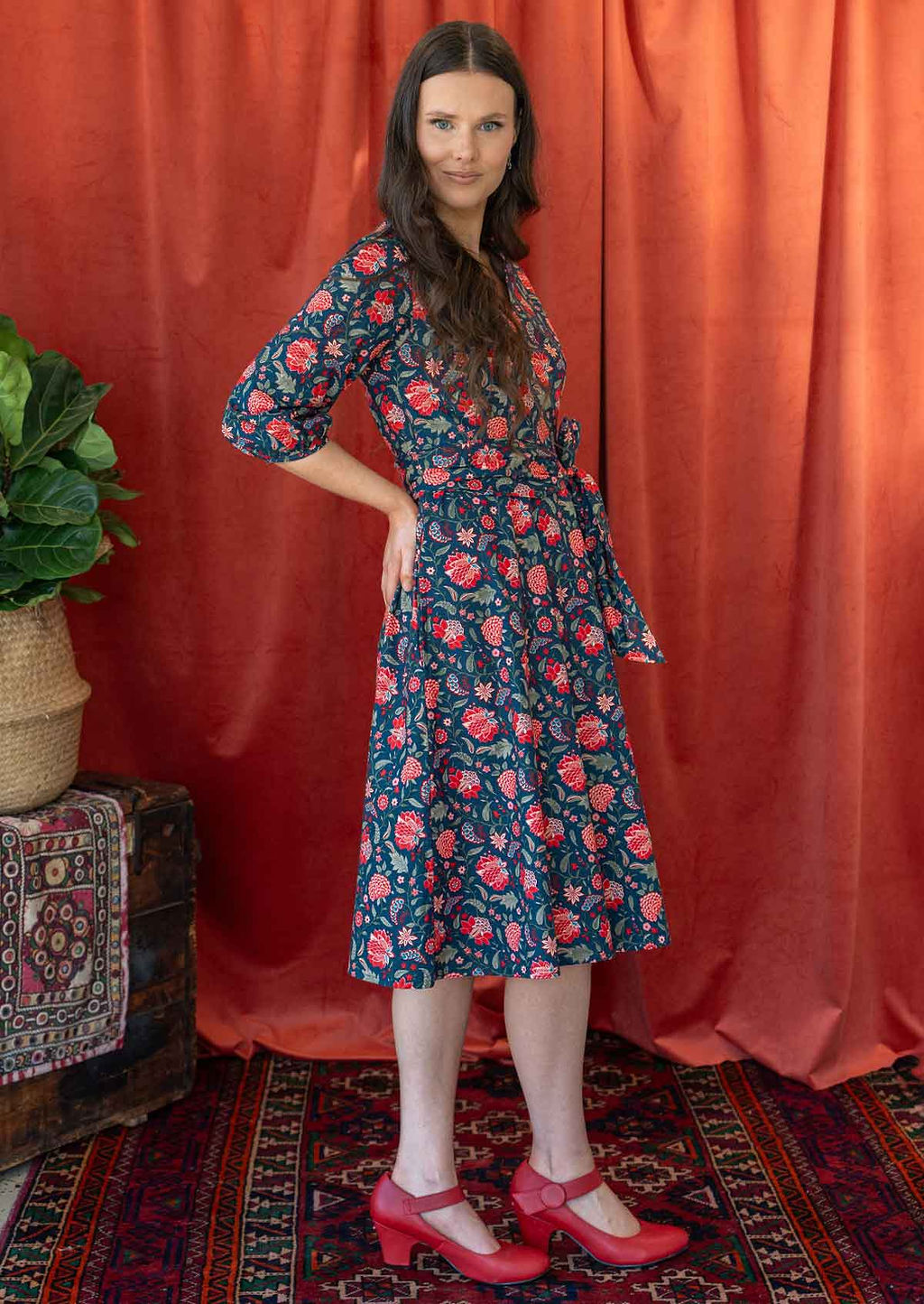 Woman stands side on in over the knee length cotton floral print wrap dress with waist-tie, with her hand on hip