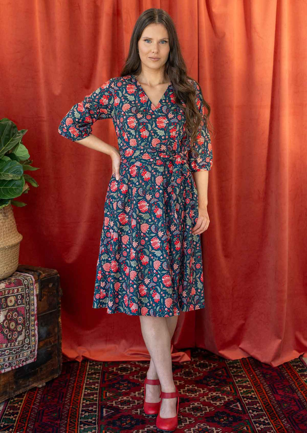 Woman wears blue based, red and green floral print cotton wrap dress with a V-neckline and a wide waist-tie