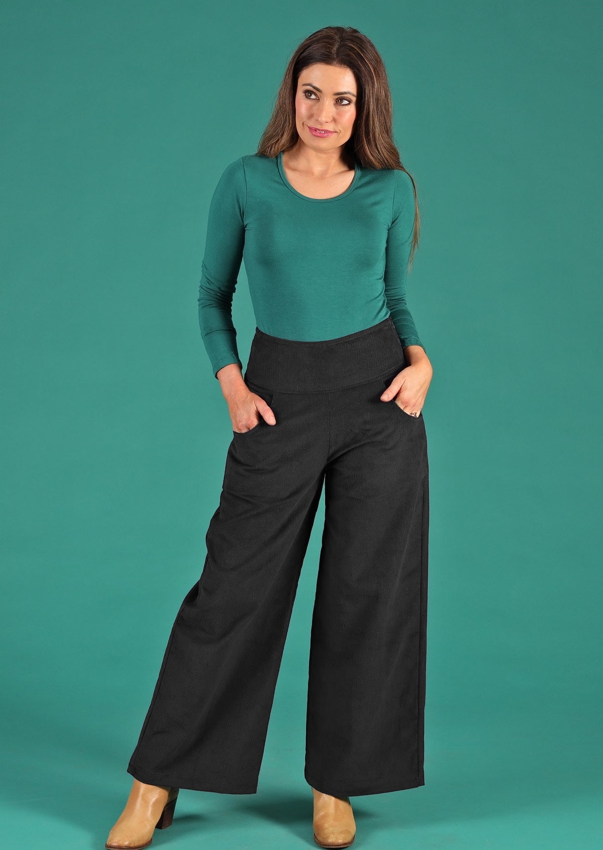 Woman stands in black corduroy wide leg pants with wide waistband, with her hands in pockets