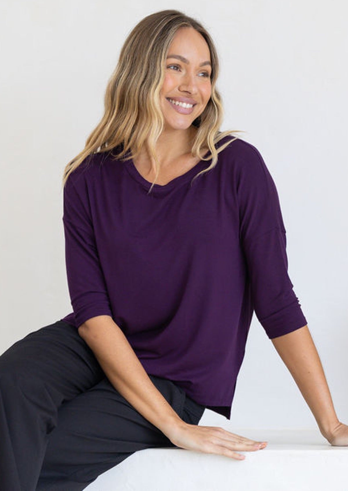 Woman sitting wearing a purple relaxed fit top with 3/4 sleeves and black pants
