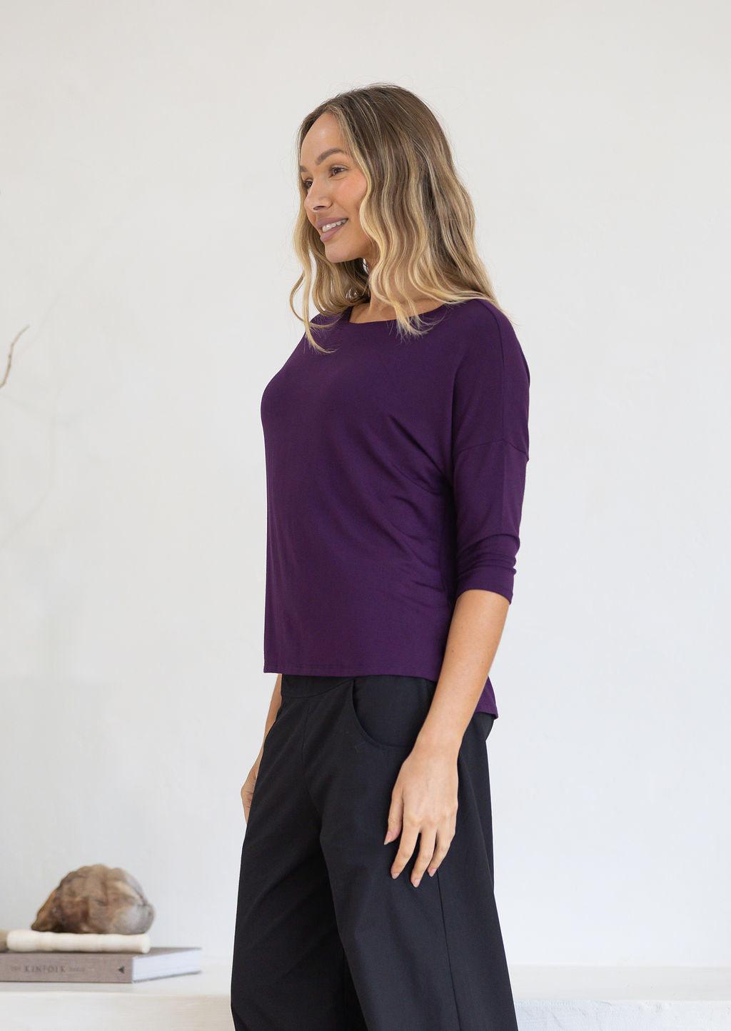 Woman wearing a purple top with 3/4 sleeves and dark pants against a white background