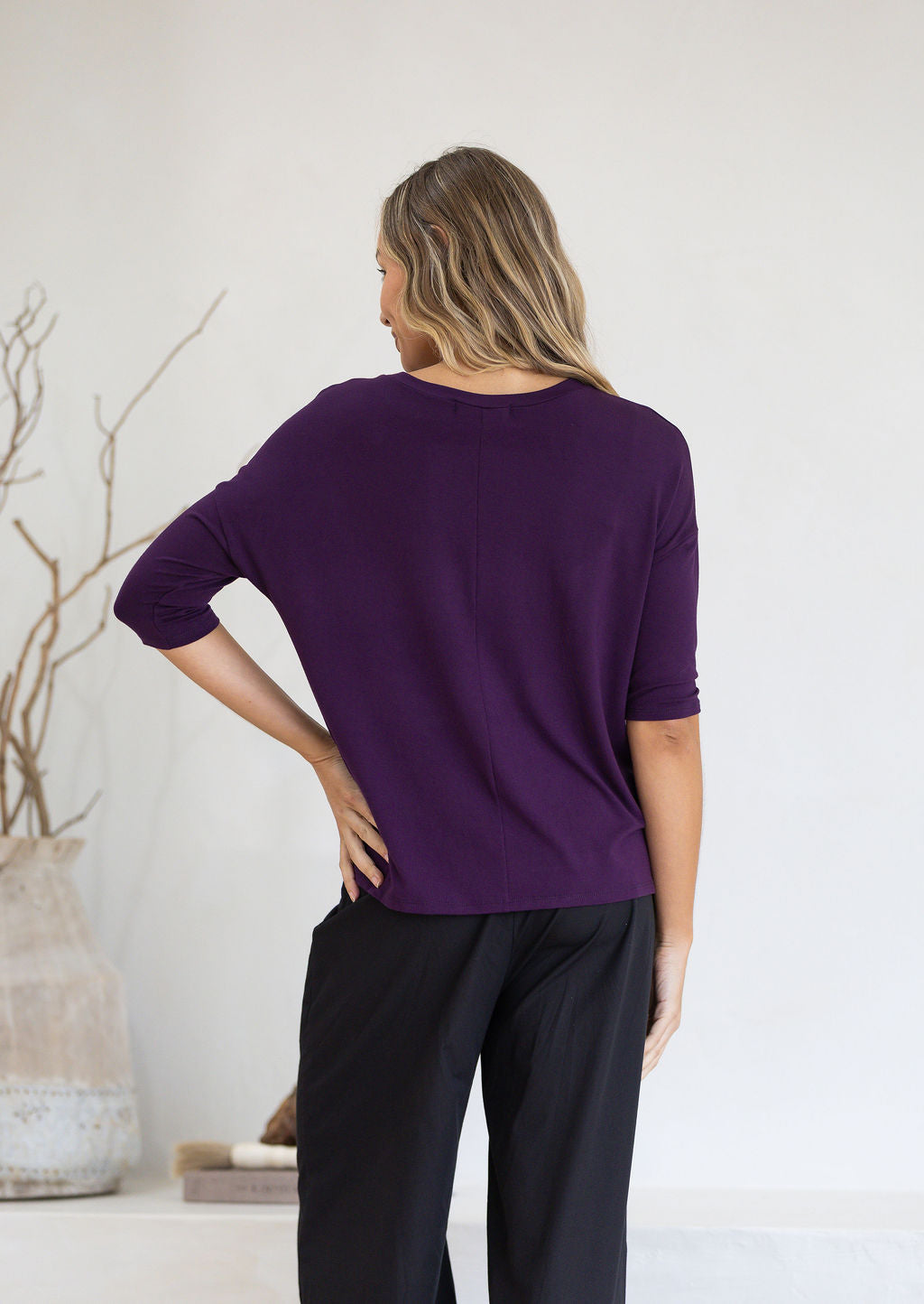 Back view of woman wearing a purple top and black pants with hand on hip