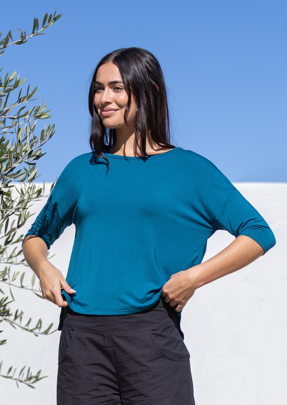 Woman holding the front hem of a teal relaxed fit top with 3/4 sleeves and black pants