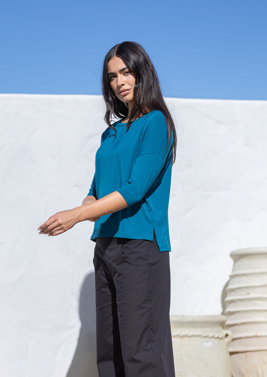 Woman wearing a teal 3/4 sleeve rayon top and black pants standing against a white wall with blue sky.
