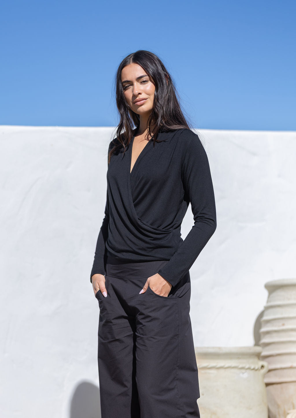 Woman wearing a black rayon false wrap top paired with black cotton pants standing against a white wall and blue sky.