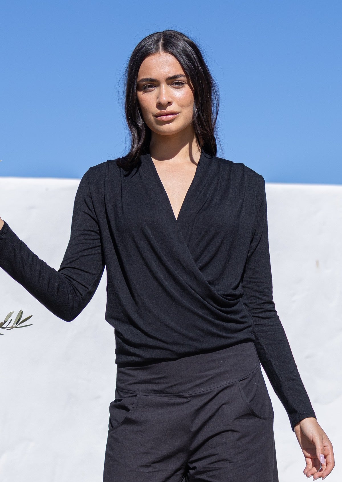 Woman wearing a black long sleeve false wrap top and pants against a blue sky and white wall.