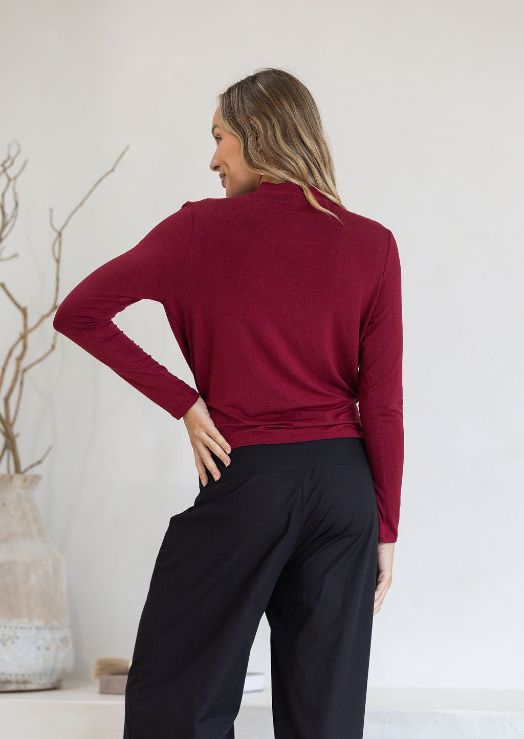 Back view of woman wearing a maroon long-sleeve top and black pants against a white background