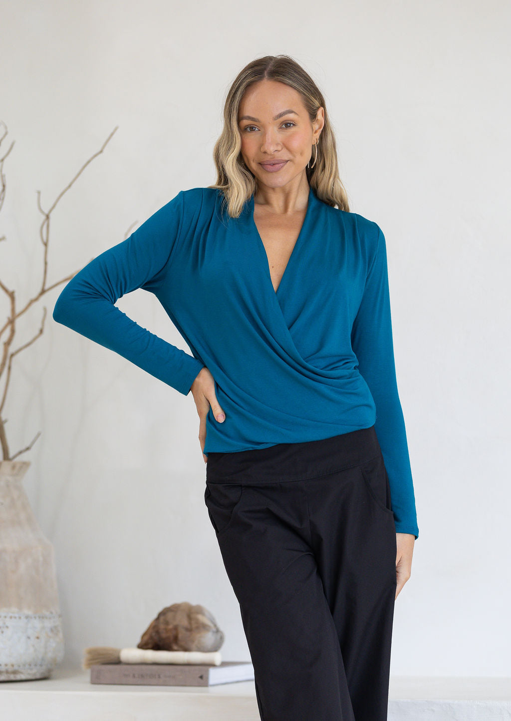 Woman wearing a teal long sleeve crossover top and black pants with hand on hip