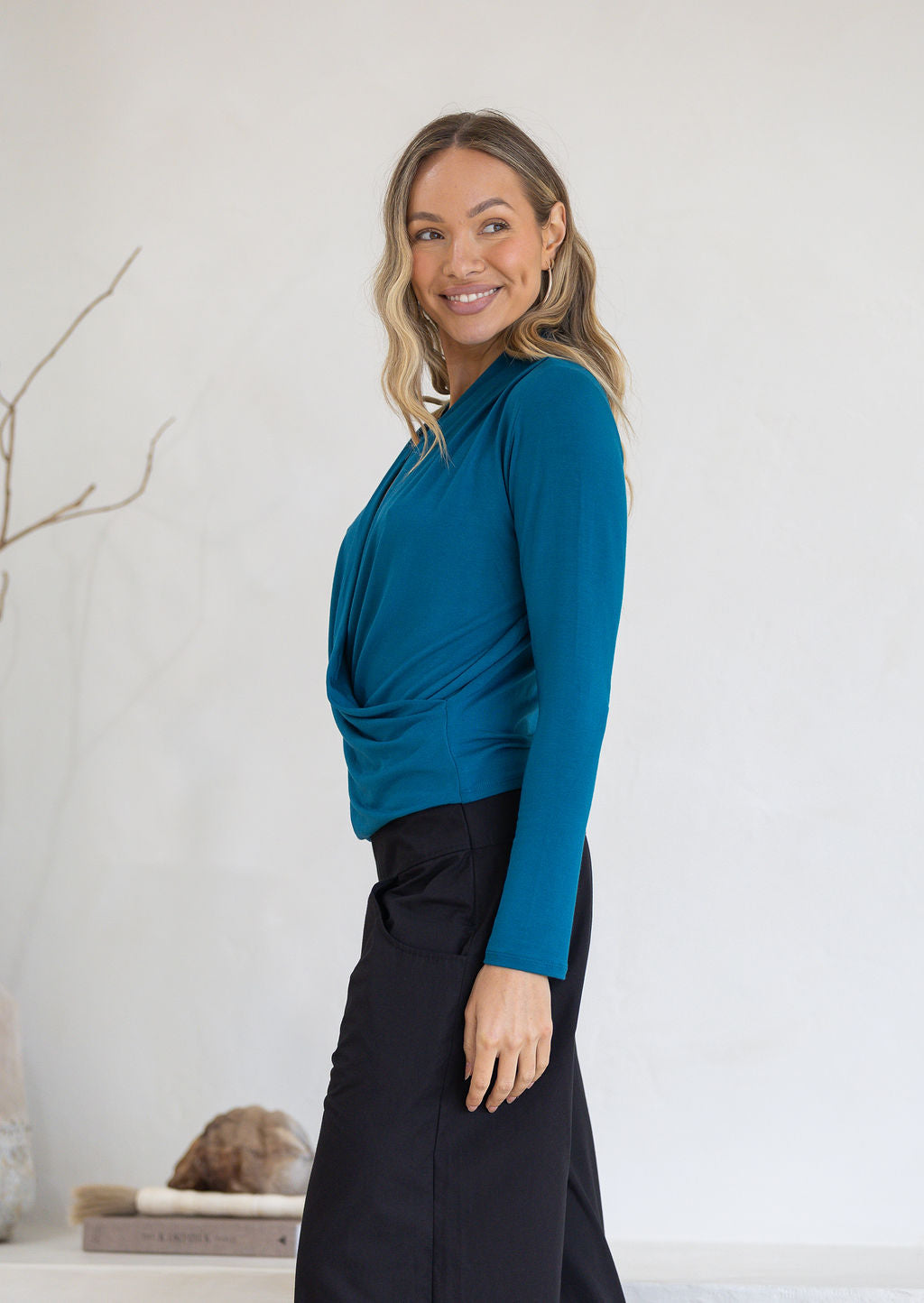 Woman wearing a teal long sleeve top with gathers where the top crosses over at the front and black pants