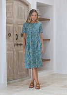 Woman wears relaxed fit cotton dress with short sleeve, pockets and small pin tucks in sage green floral print.