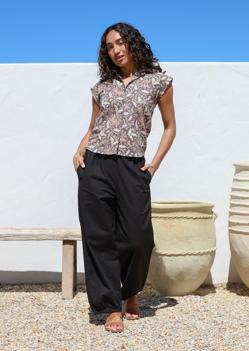 Woman with hands in pocket wears wide leg black cotton pants with elastic at either side of waist and pleated ankles