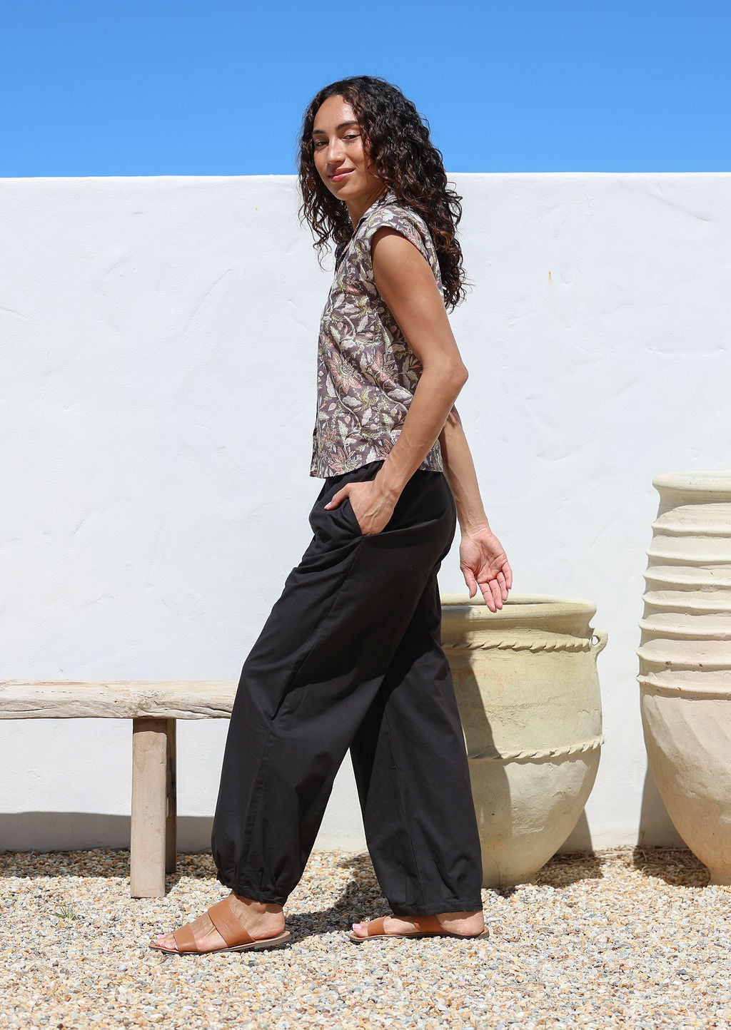 Woman in a floral shirt top and black wide leg cotton pants standing outdoors with a white wall and blue sky in the background