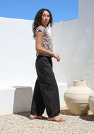 Model stands in relaxed fit black cotton pants with elasticated waist, tapered ankles and side pockets.